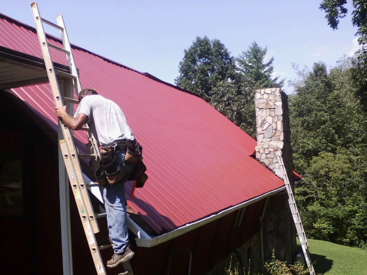 Roofer on a ladder beside a red metal roof and stone chimney — Soffit & Fascia Repair by Roofing Brothers in Waldport